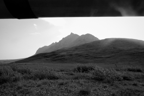 A black and white photo of a section of the Trans-Alaska Oil Pipeline in the foreground -- underneath it, permafrost tundra stretches across vast hills. In the background, peaks of the Brooks Range, within the Arctic National Wildlife Refuge, extend into a white sky.