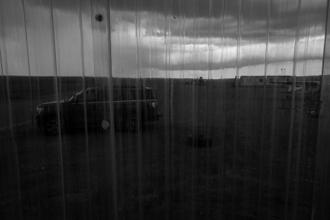 A black and white photo, taken through a transparent window pane from a USFWS bunkhouse porch. In the foreground, seen through the clear panels, a black SUV sits on a gravel airstrip. In the background, dark storm clouds hover.