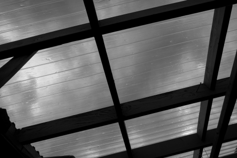 A black and white close-up of the USFWS bunkhouse's transparent porch roof. The square, clear panels are held by criss-crossed wooden beams which. Raindrops are visible through the roof, sliding down their slanted panes.