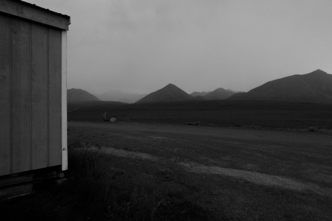 A black and white photo of storm clouds rolling over the Brooks Range, their peaks in the background being engulfed by haze and rain. In the foreground, the corner of a wooden bunkhouse extends into frame from the left. Tundra grass and gravel extend to the base of the mountains.