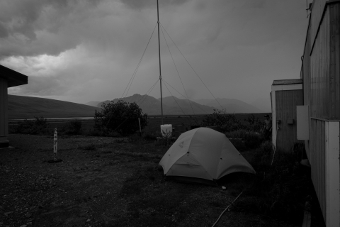 A black and white photo of a tent pitched between two rectangular buildings (U.S. Fish and Wildlife bunkhouses), both just barely in the shot. In the background, the Brooks Range is hazy as moving rainclouds make their visibility hazy.