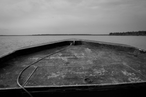A black and white photo of the nose of a motor boat, taken from within the boat, as it rumbles through the Yukon River. Beyond the boat, and everywhere around it, is water.