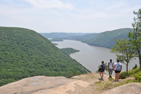 Three people stand on a rocky ledge overlooking the Hudson River