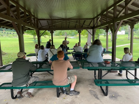 A group of people sitting at a picnic tables listening to a presentation.
