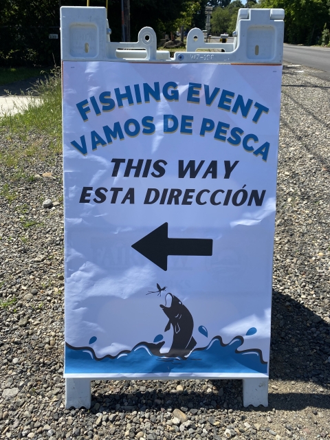 Sign pointing left that reads "fishing event, this way" and "vamos de pesca, esta direccion" with a drawing of a fish jumping out of the water to catch an insect.