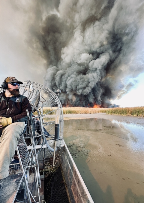 A firefighter sits in an airboat with a large plume of smoke in the background.