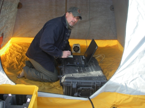 A man in a jacket and cap kneels over a large black machine inside a white and yellow tent. 