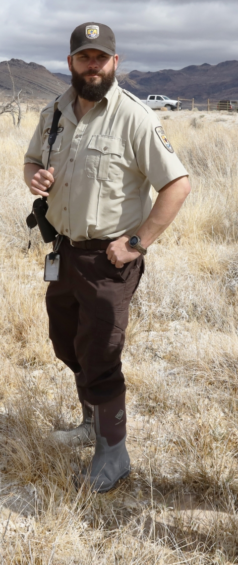 Man in brown pants and hat with U.S. Fish and Wildlife Service shield on desert-like landscape with tan grasses and mountains and white truck visible in the background.