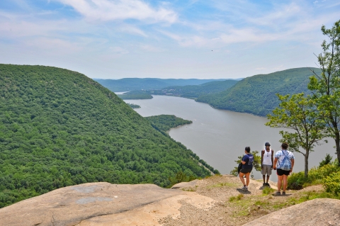 Three people stand on a rocky ledge overlooking the Hudson River