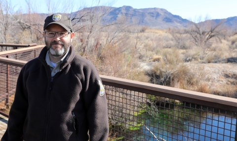 Man with brown jacket and hat with U.S. Fish and Wildlife Service shield stands in front of body of water with mountains and dry desert landscape with trees in the background.