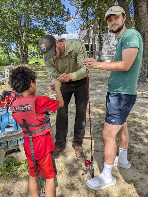 a person in USFWS uniform helps a kid and a young man attach a lure to their fishing rod