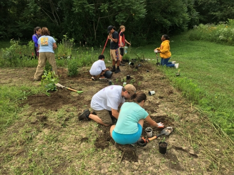 Two adults work with six middle school-aged children to remove plants from pots and place them in the prepared soil of a new pollinator garden. 