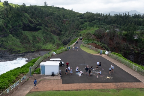 Visitors on a large black walkway at top a bluff are walking and making chalk art. The walk way is surrounded by lush green vegetation and an ocean cove can be seen on the left side. 