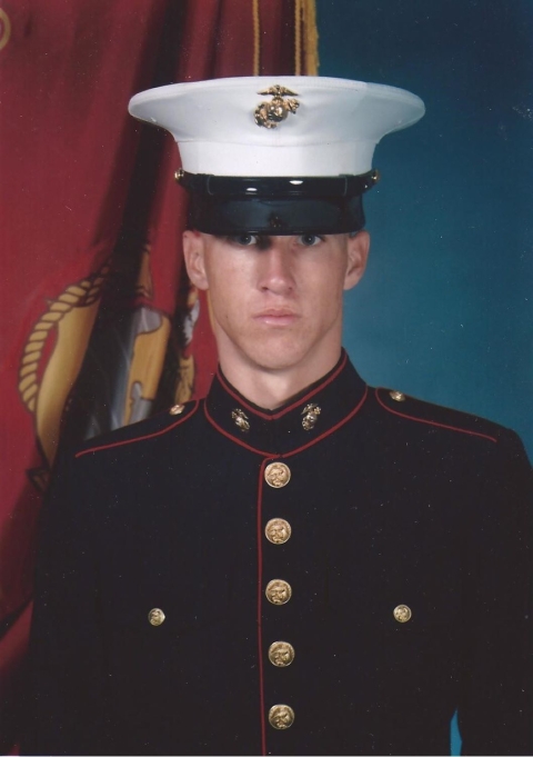 A formal portrait of a man wearing a dark blue Marine Corps dress uniform with gold buttons and a white hat posed in front of the Marine Corps flag