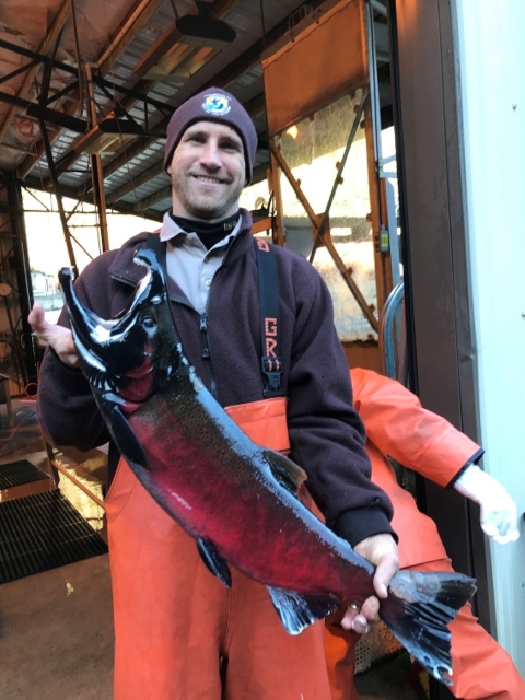 A man smiles big while holding a very big fish. He wears orange waders and a brown jacket and beanie.