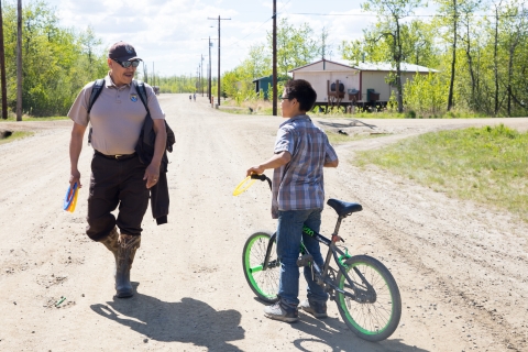 A Refuge Information Technician in uniform walking and talking to a young kid on a bike on a dirt village road while the kid holds a bicycle. 