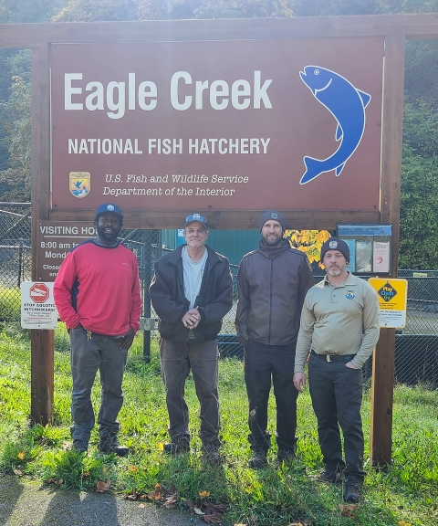 Four men stand smiling in front of a large sign for the Eagle Creek National Fish Hatchery. Sunlight and green grass are behind them.