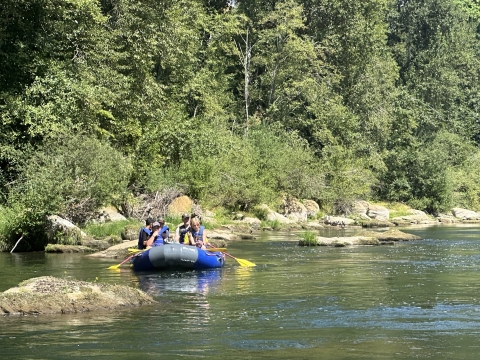 A raft with a few people and oars in the water float in a calm river with a backdrop of green trees.
