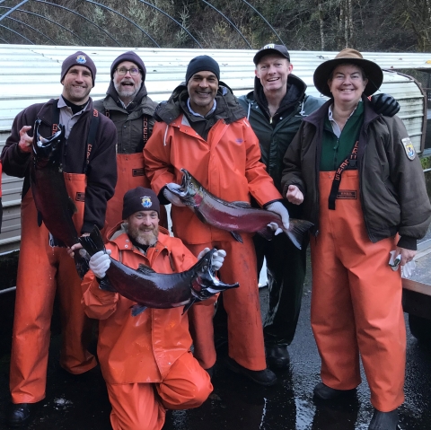 A group of six people all smiling and wearing organge waders. Three of them are holding large fish.