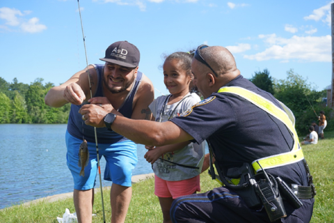 a policeman and man hold up a fishing rod with a fish on the line while a young girl smiles in excitement