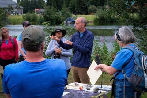 Owen Guthrie stands within a group of FWS employees, partners, and members of the local media, explaining the history of Cripple Creek. In the background, before stalks of fireweed, the Chena River flows. 