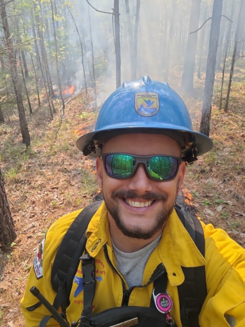 Nathan Herron smiles at the camera while in fire gear in the woods
