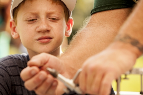 A boy looks intently at the hands of an adult holding or making a tool.