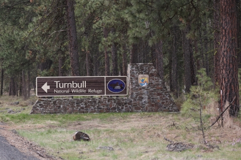 Wooden sign with text that reads "Turnbull National Wildlife Refuge" and an arrow pointing to the left. A small portrait of a redhead duck is also on the sign.
