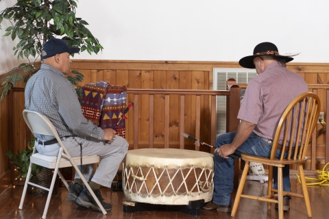 two men sit in chairs while hitting a large drum placed between them