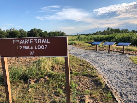 A brown sign reads "Prairie Trail" half mile loop with a hiker icon. The sign is next to a gravel trail. There are green trees and plants in the background. 