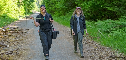 Two Service interns walking down a wooded path. The intern on the left is holding a radio telemetry tracking antenna and receiver.
