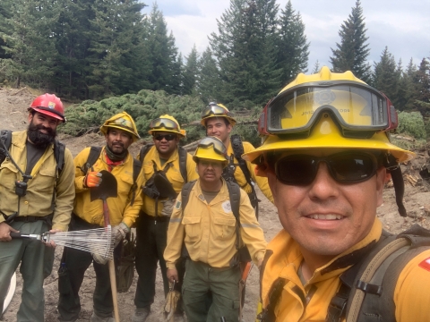 A firefighter takes a selfie with a group of other firefighters behind him. They are all wearing their firefighting gear and helmets.