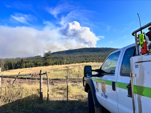 A view of a USFWS fire engine in front of a fence and field. In the background is a wildfire clearly visible on the hillside.