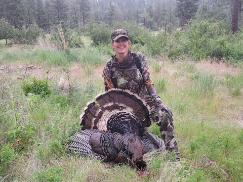 A woman wearing camouflage kneels beneath a turkey she harvested