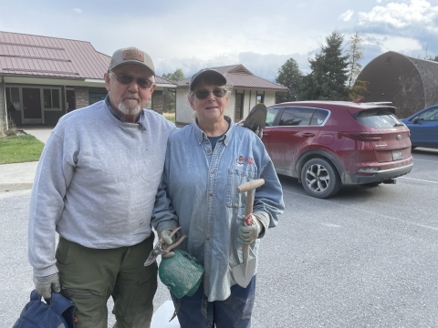 Two volunteers smile while standing in a parking lot. Both hold tools in their hands