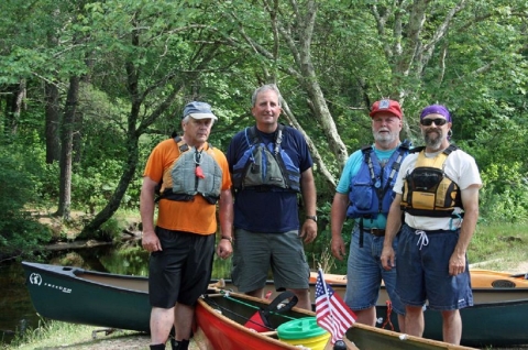 Four white men wearing life vests stand next to canoes on a river bank