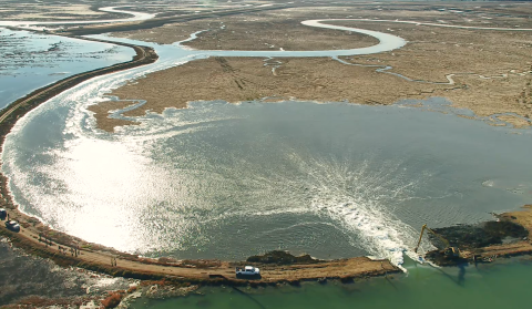 Aerial photo shows digging machine breaking earthen barrier and allowing bay water to flow into pond.