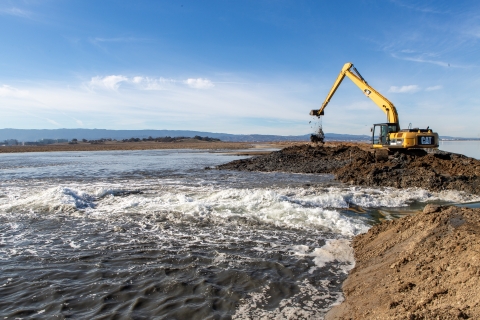 A bulldozer works to remove part of an earthen levee separating two bodies of water. 