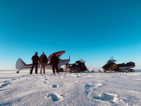 Three people stand on a flat snow covered landscape with a bush plane and two snow machines.