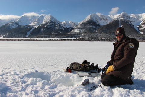 A person kneels in the snow on a frozen lake with mountains in the background.