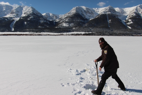 A person digs a hole in the snow on a frozen lake with mountains in the background.