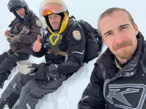 Three Federal Wildlife Officers sitting on snow wearing snow machine personal protective gear. 