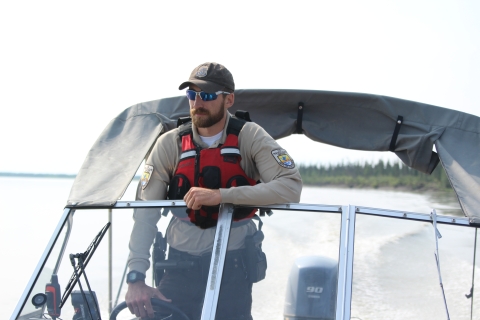 A view from the bow of a boat towards the stern of a Federal Wildlife Officer at the helm with the Yukon River in the background. 
