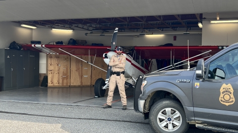 Federal Wildlife Officer Cody Smith stands inside a hangar in front of a Top Cub bush plane with his patrol truck parked in front of the open hangar door. 