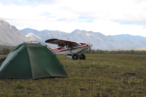 A small tent with a Top Cub bush plane parked behind it on a grass runway area with mountains in the background. 