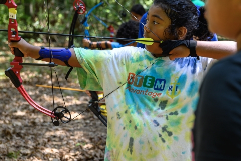 Inidgenous youth pulling arrow on bow and arrow, about to shoot, wearing a "STEM Advantag RI" t-shirt. 