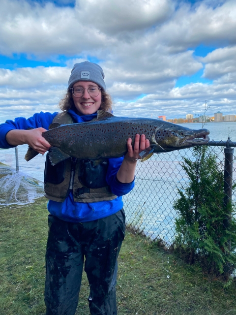 A woman wearing a gray beanie, glasses, and blue jacket smiles while holding up a large fish. Clouds, blue sky, and water, are behind her.