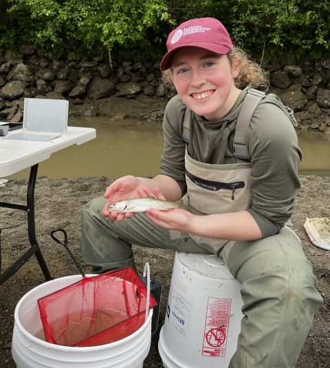 Service intern, Kathleen Gerard, holds a coastal cutthroat trout while seated. Fish sampling buckets and equipment are in view around her and water and vegetation are in the background. 