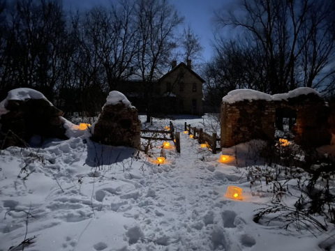 Snow-covered ground is complemented by lit up ice luminaries on alternating sides of a snow-covered path, going thorugh brick barn ruins with picket fence on each side. A brick house can be seen in the background through trees and a purple night sky.