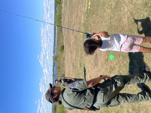 Officer smiling as a young girl casts a fishing rod on a green lawn. 